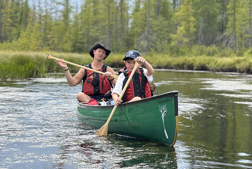 A man and young boy canoeing on a calm river surrounded by trees, both wearing life jackets and paddling with wooden oars.