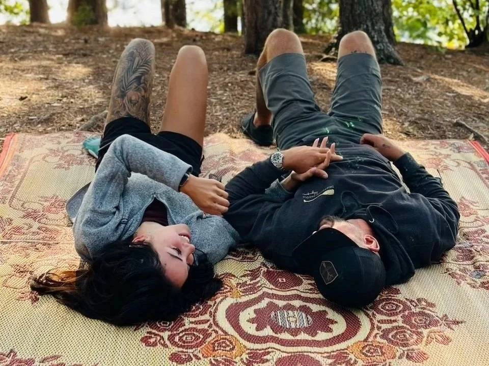 A couple lies on a patterned outdoor rug on the ground in a wooded area, holding hands and looking at each other.
