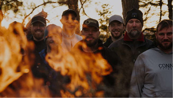 Group of men smiling around a campfire outdoors at sunset, with trees and a clear sky in the background.