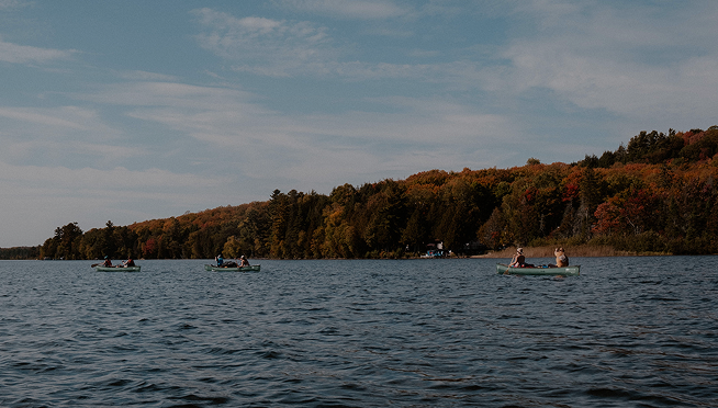 People kayaking on a lake with a tree-covered shoreline and colorful fall foliage under a partly cloudy sky.