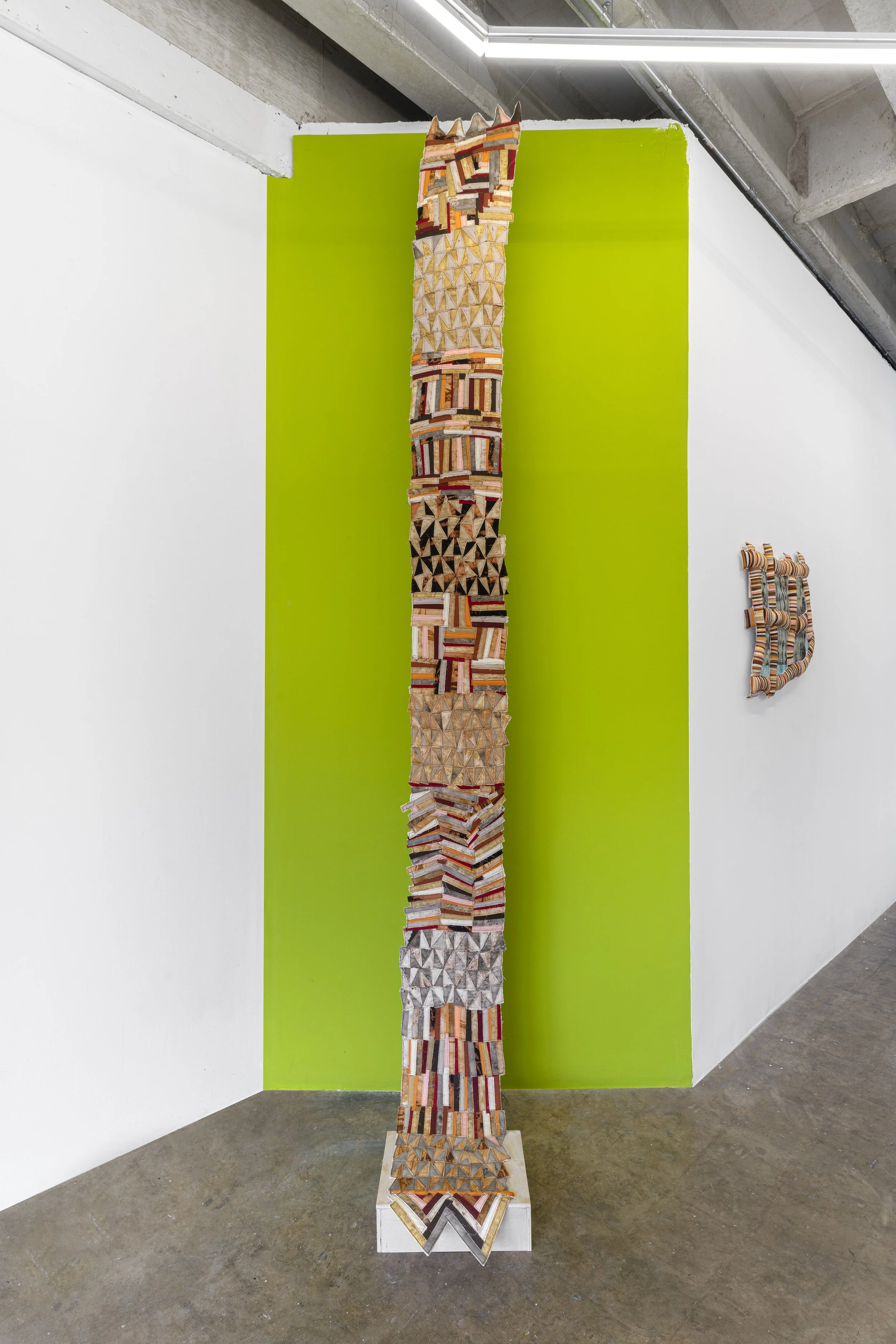 Tall abstract textile artwork with geometric patterns, placed on a white base against a bright lime green wall in an art gallery.