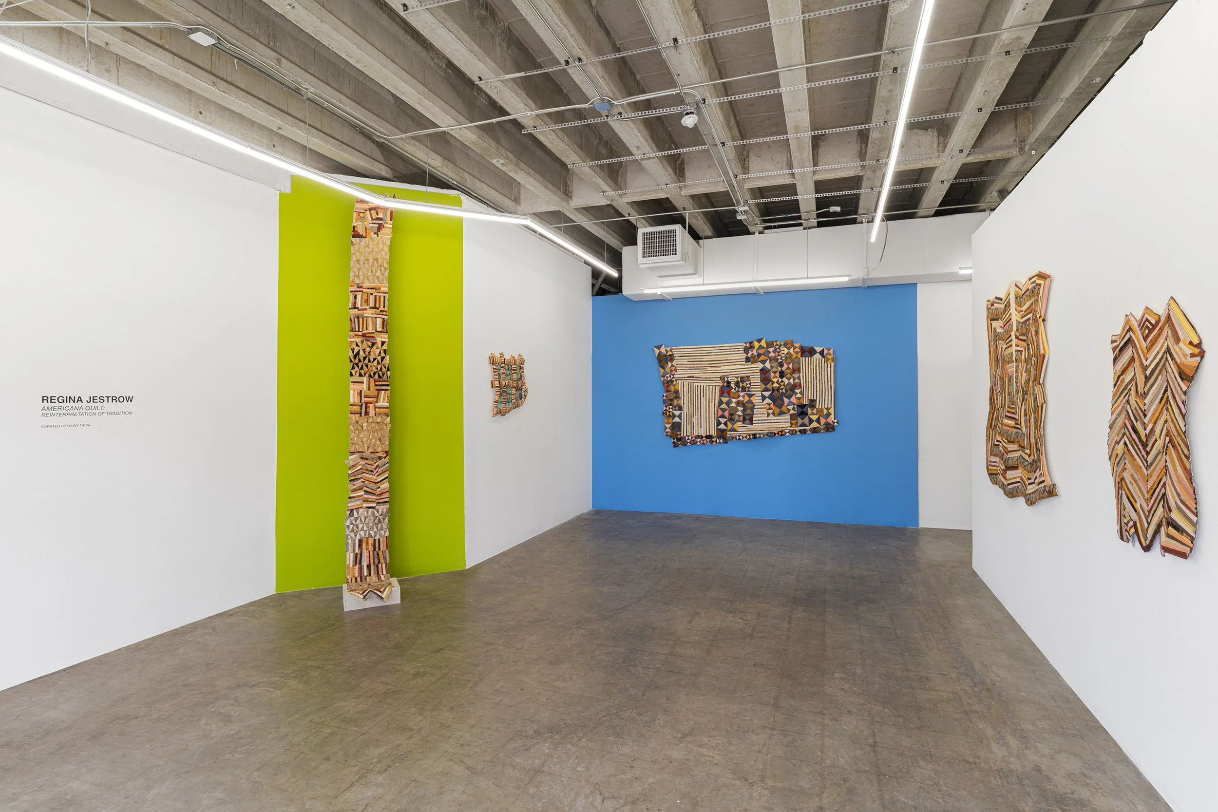 Art installation titled 'American Quilt' by Regina Jestrow featuring various colorful quilt artworks mounted on white and blue gallery walls with a gray concrete floor.