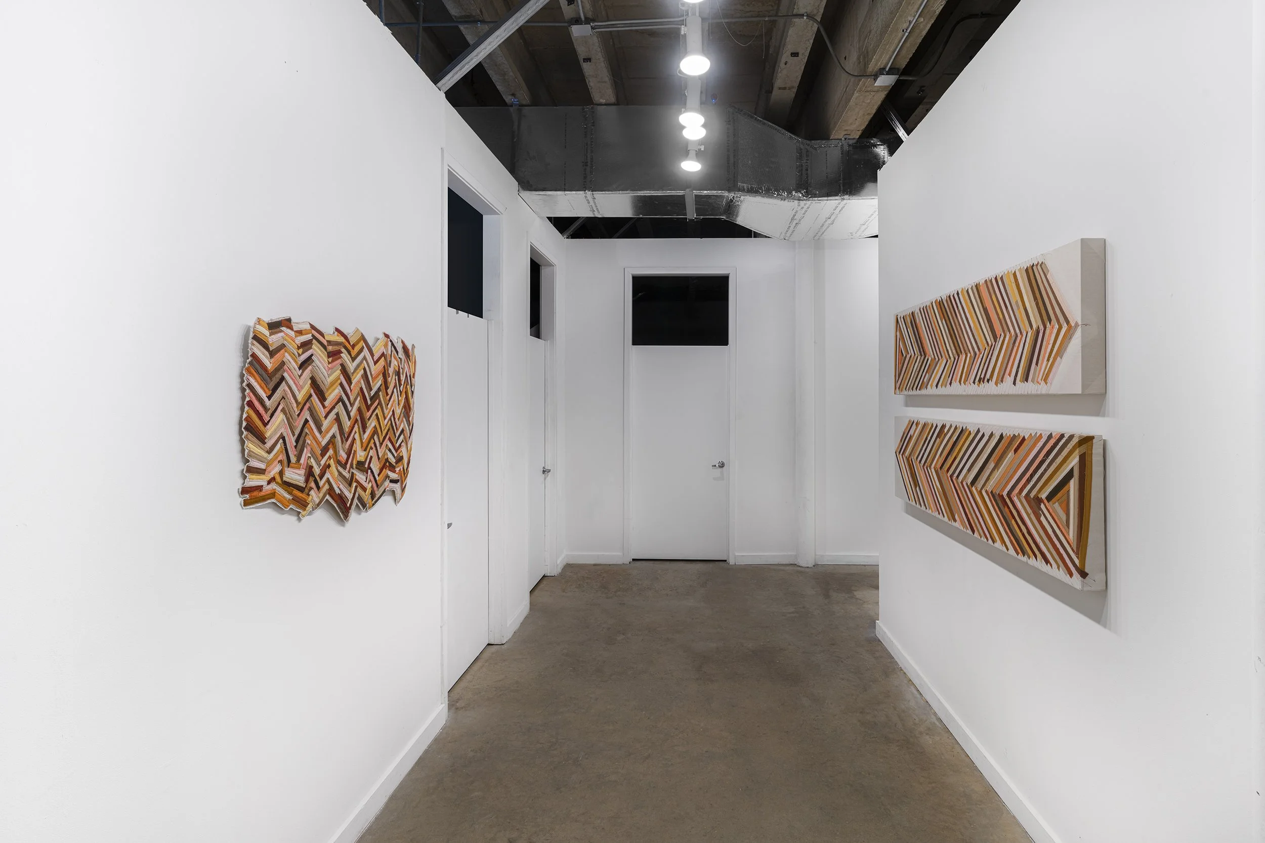 Art gallery with white walls, featuring two textile or mixed media zigzag patterned artworks on the left and two similar artworks on the right, with a concrete floor and an industrial ceiling.