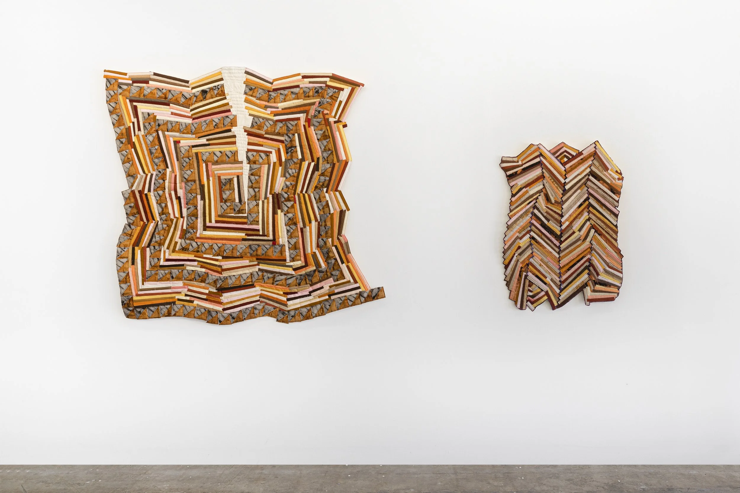 Two wall-mounted art pieces made of layered, colored pencil tips arranged in geometric patterns on a white gallery wall.