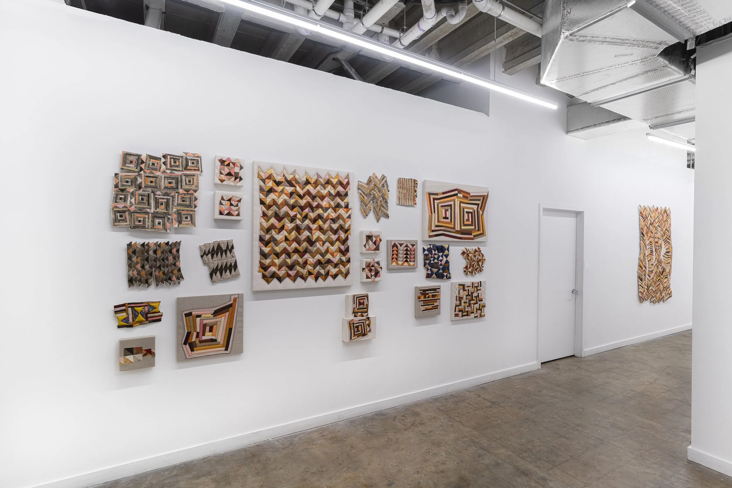 Gallery wall with various geometric patterned textile art pieces in a modern art gallery with white walls and industrial ceiling.