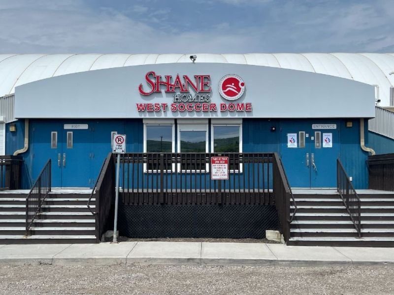Front view of Shane Homes West Soccer Dome, with blue exterior walls, stairs leading to the entrance, and signage indicating entry and exit.