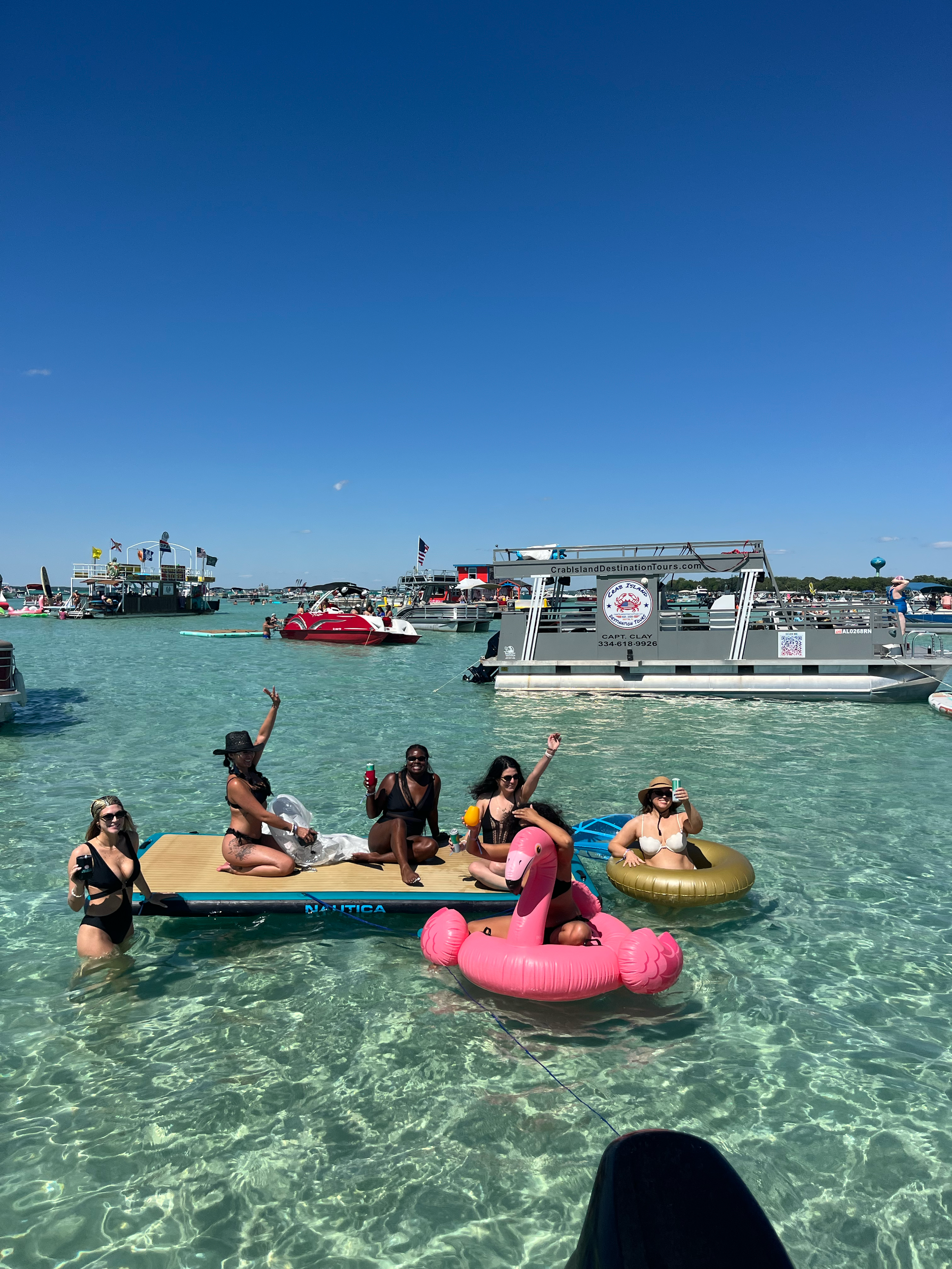 Group of people enjoying the water on inflatables, including a pink flamingo float, with boats and clear blue sky in the background.