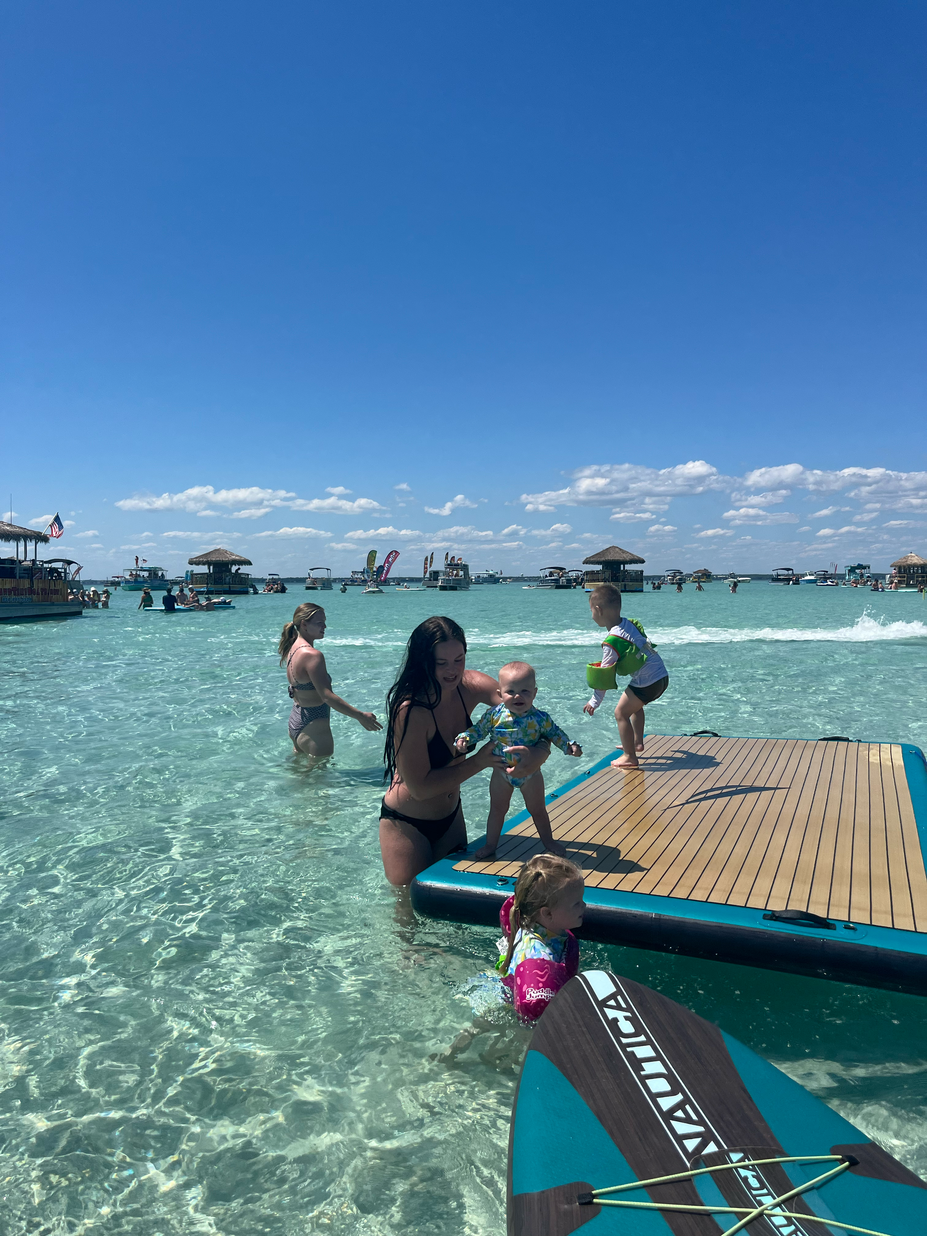 People enjoying a day at the beach with a paddleboard and inflatable platform on clear, shallow water under a blue sky.