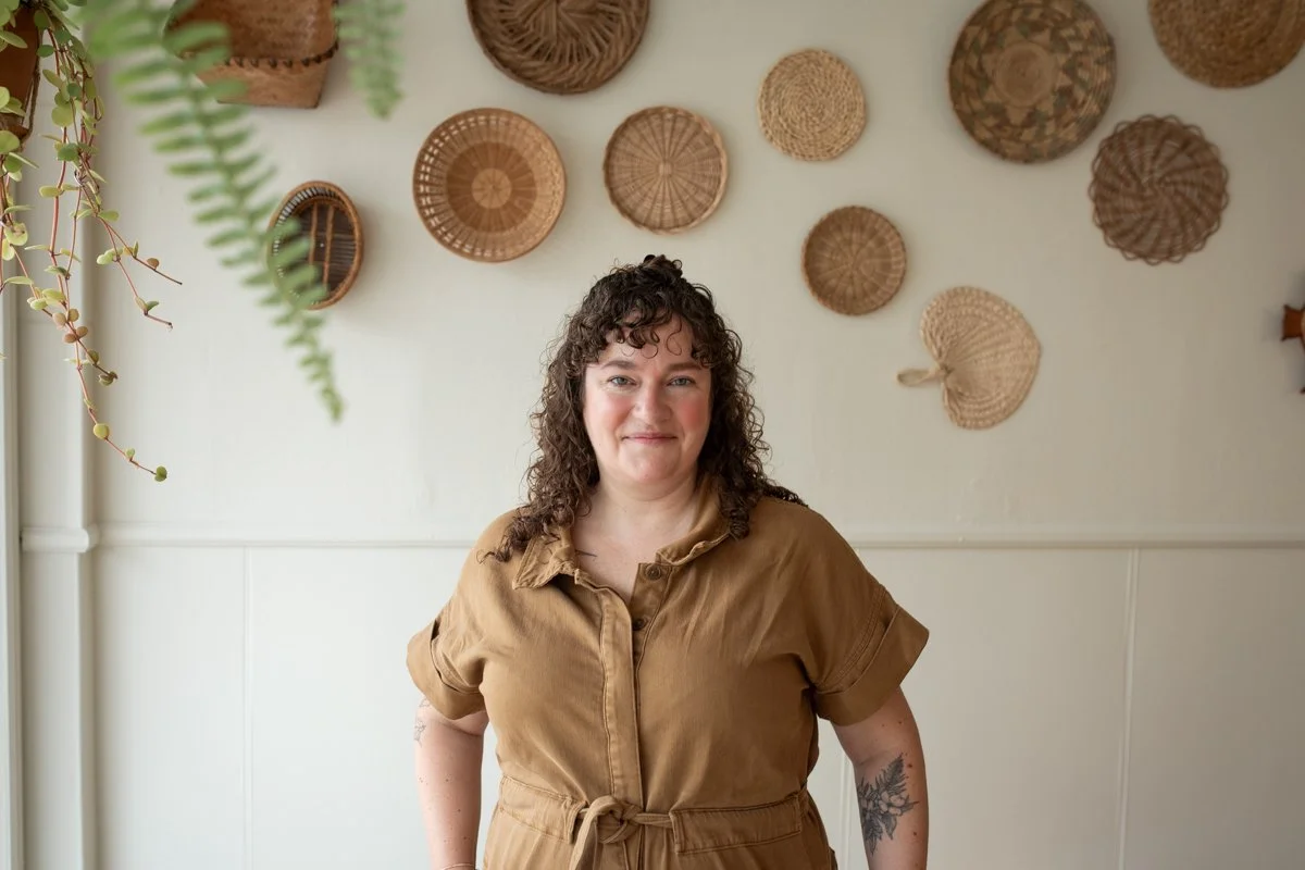 Smiling woman in a brown jumpsuit with baskets hung on a wall behind her in Portland, Oregon
