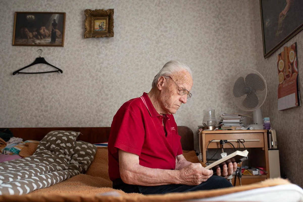 An elderly man with gray hair and glasses, wearing a red polo shirt, sitting on a bed and reading a book in a bedroom