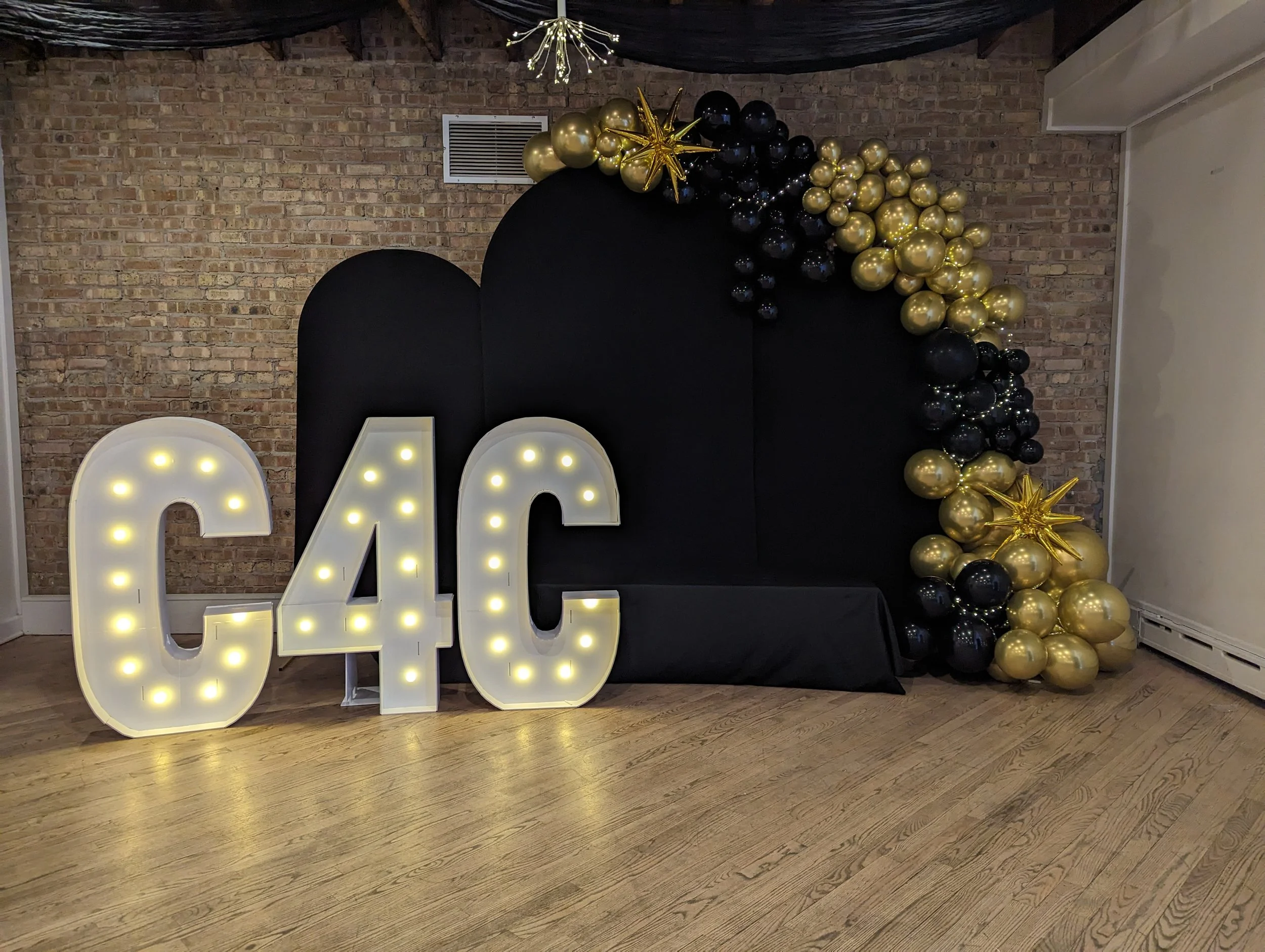 Decorative setup with large illuminated letters 'C4C' on the floor, with black and gold balloons arranged in a semi-circle behind a black backdrop, in front of a brick wall.