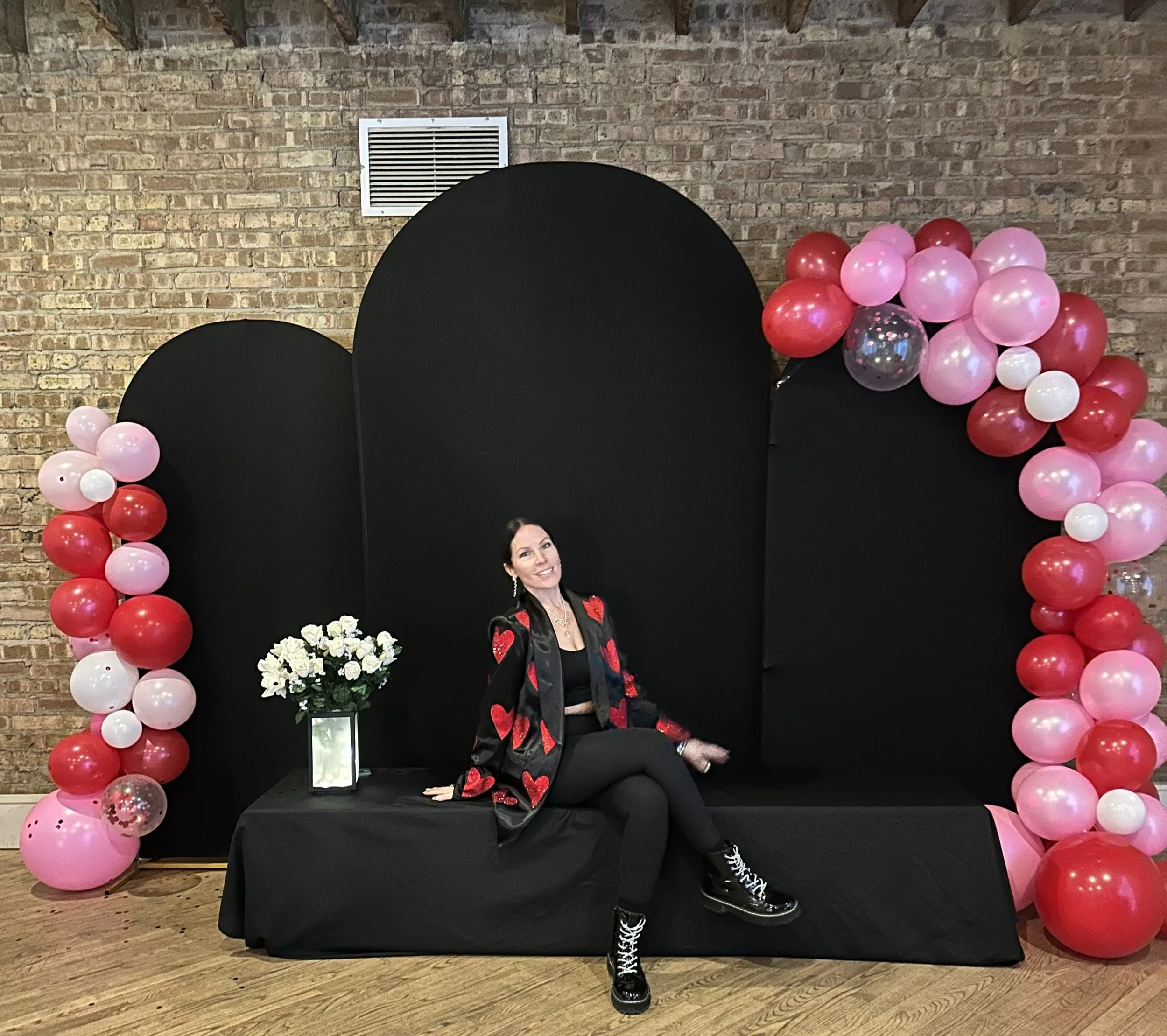 Woman sitting on a black cushion with a backdrop of black arches and festive balloon decorations in red, pink, and white, with a bouquet of white flowers nearby.