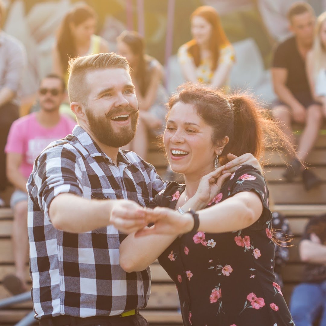 A couple dancing and smiling at an outdoor event with an audience sitting on bleachers in the background.