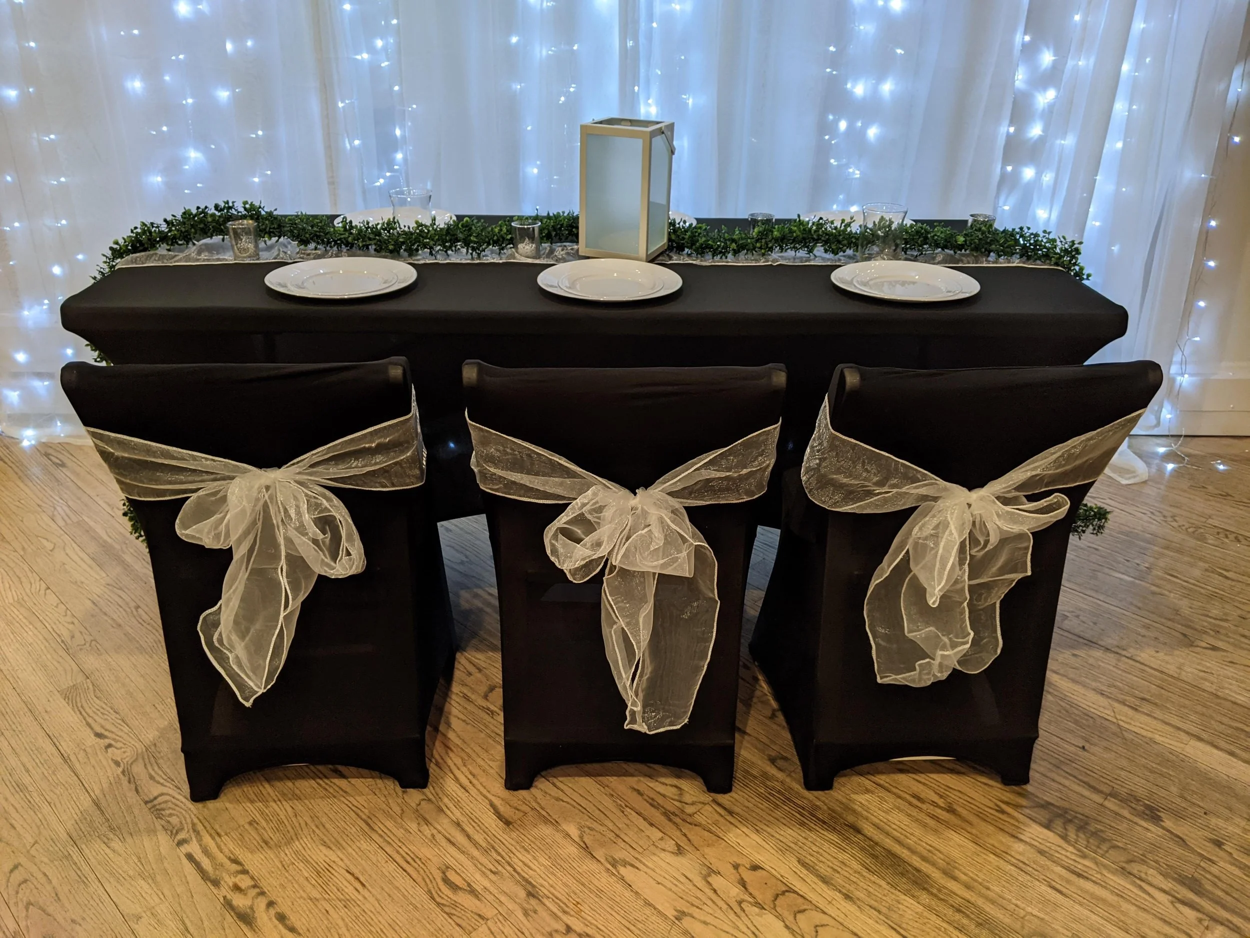 Table set for a celebration with three black chairs adorned with white ribbon bows, a black tablecloth, a centerpiece greenery runner, three plates, a lantern, and fairy lights backdrop.