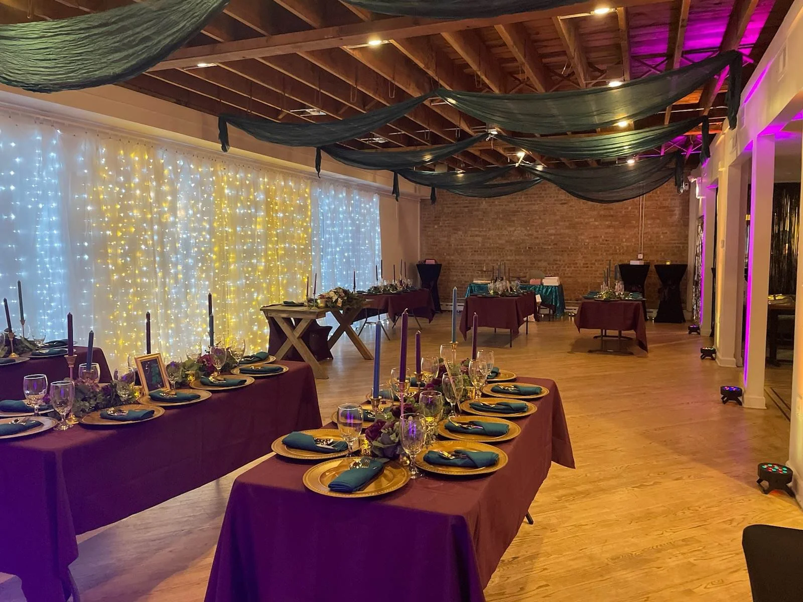 Event banquet hall decorated with purple tablecloths, candle centerpieces, and string lights with drapery on the ceiling and a brick wall in the background.