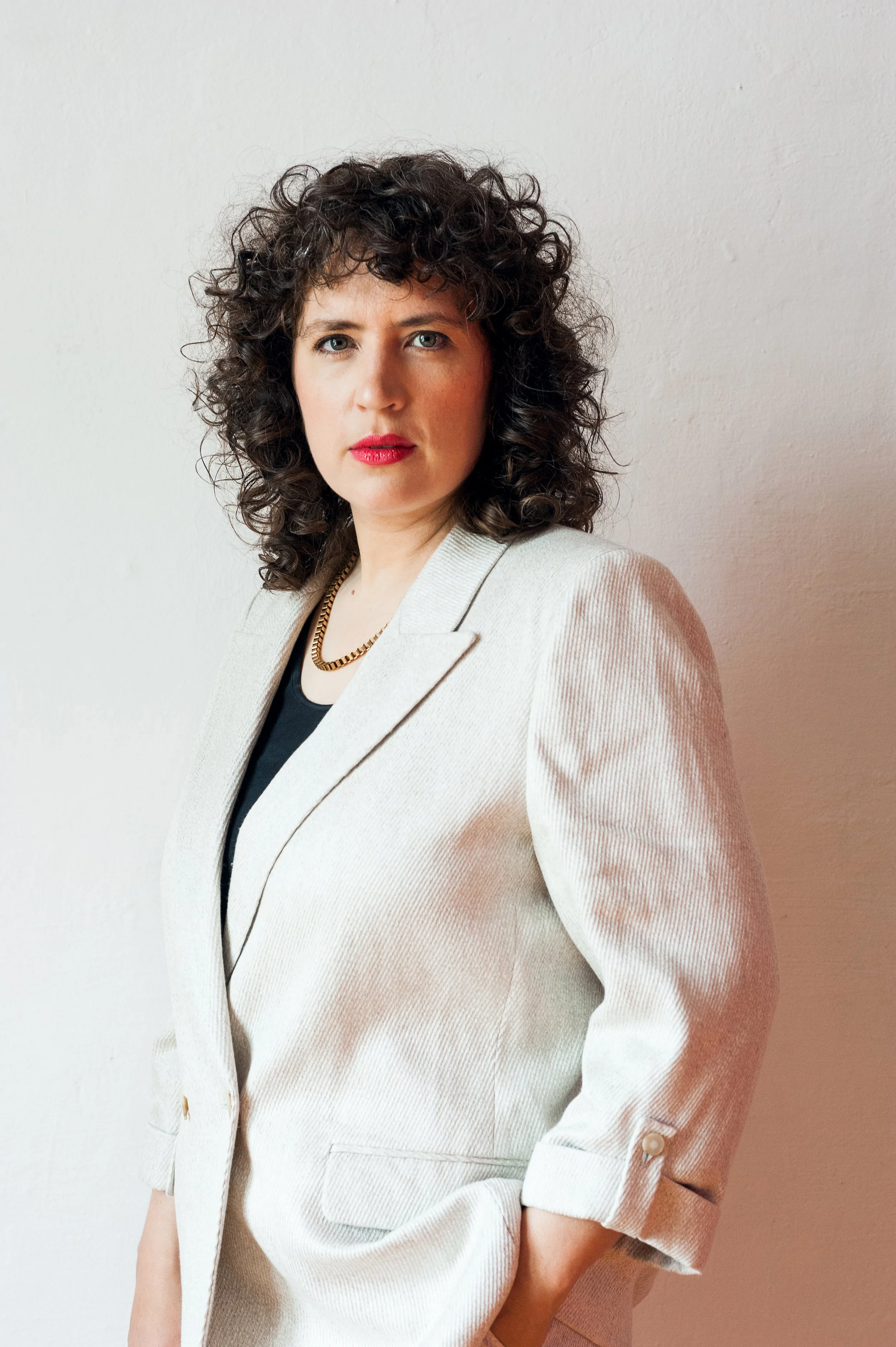 A woman with curly dark hair, wearing a white blazer over a black top, and a gold necklace, standing against a plain white wall.
