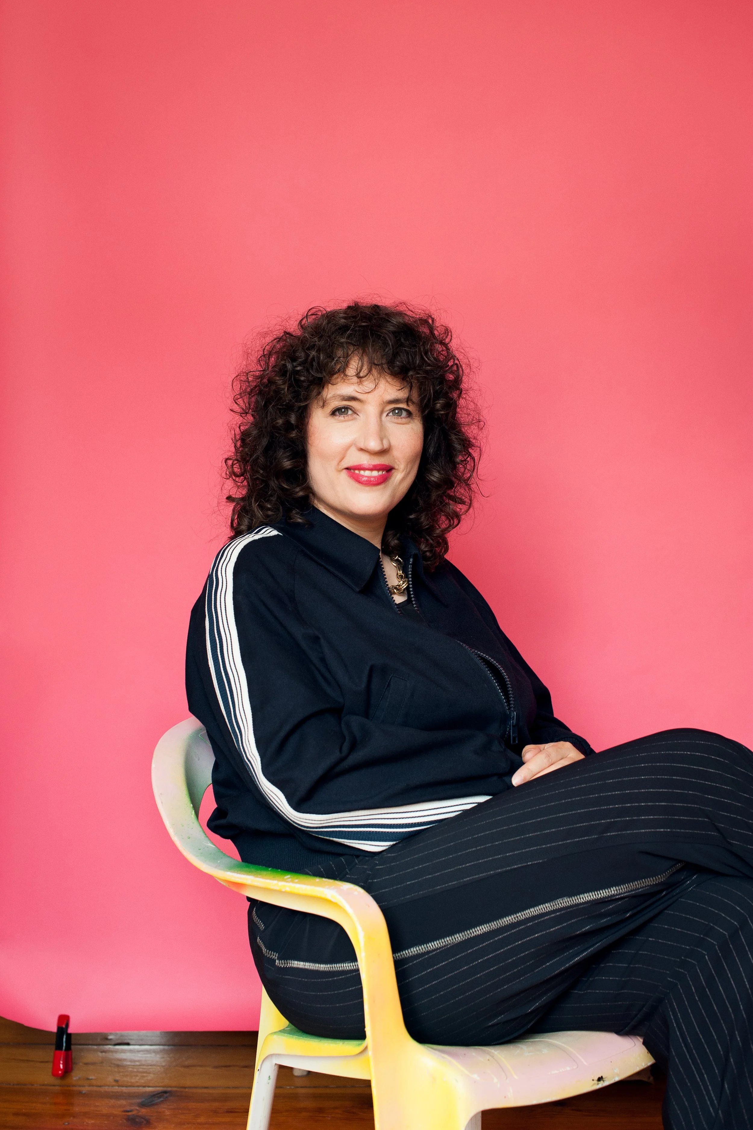 A woman with curly dark hair and light skin sitting on a colorful chair against a pink background. She is wearing a black jacket with white stripes on the sleeves and black pants with white pinstripes. She is smiling, looking at the camera, with her hands folded on her lap.