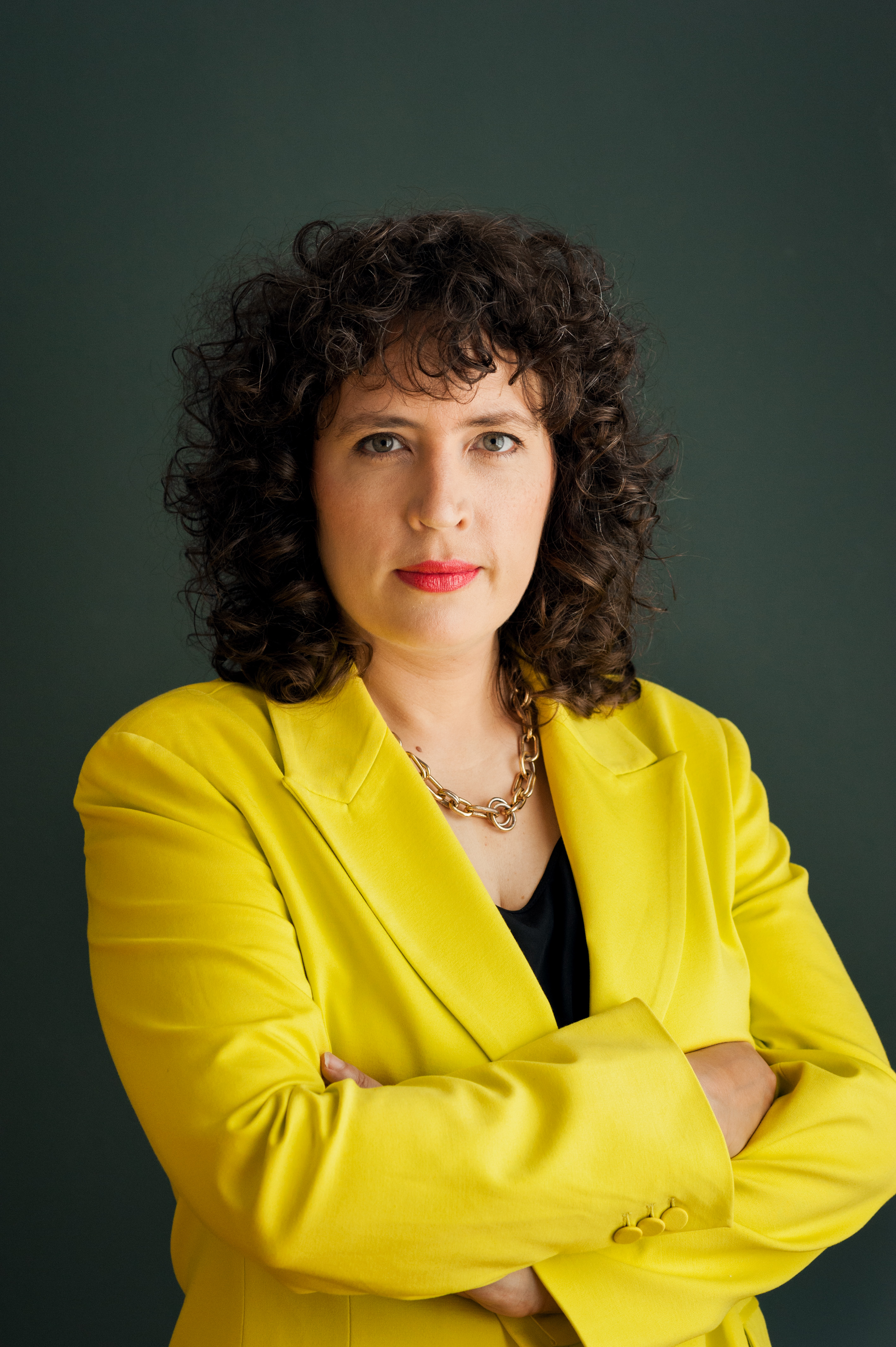A woman with curly dark hair and blue eyes, wearing a bright yellow blazer and a black top, stands with her arms crossed against a dark green background.