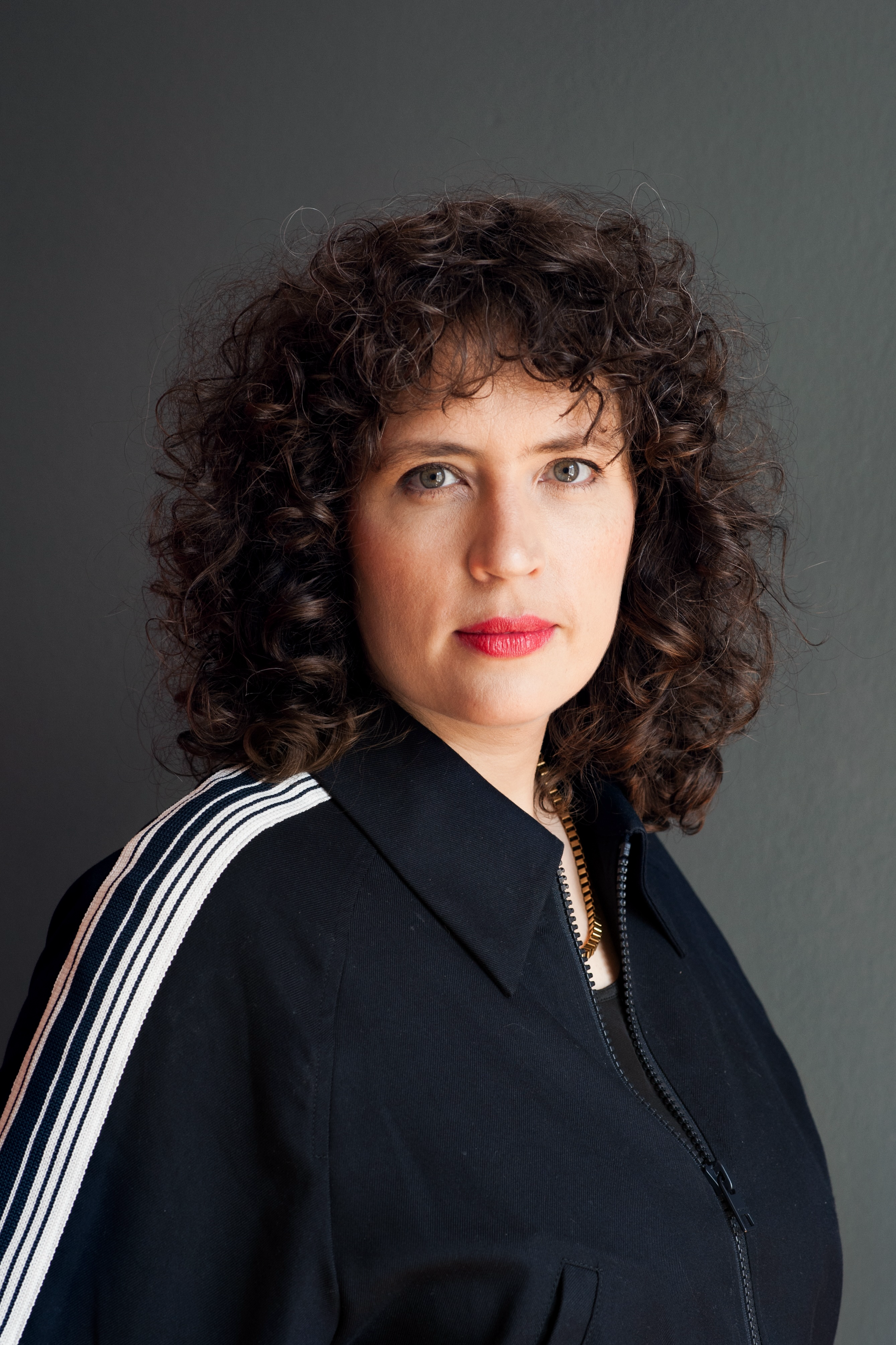 A woman with curly brown hair and fair skin wearing a black zip-up jacket with white stripes on the shoulders and a gold chain necklace.