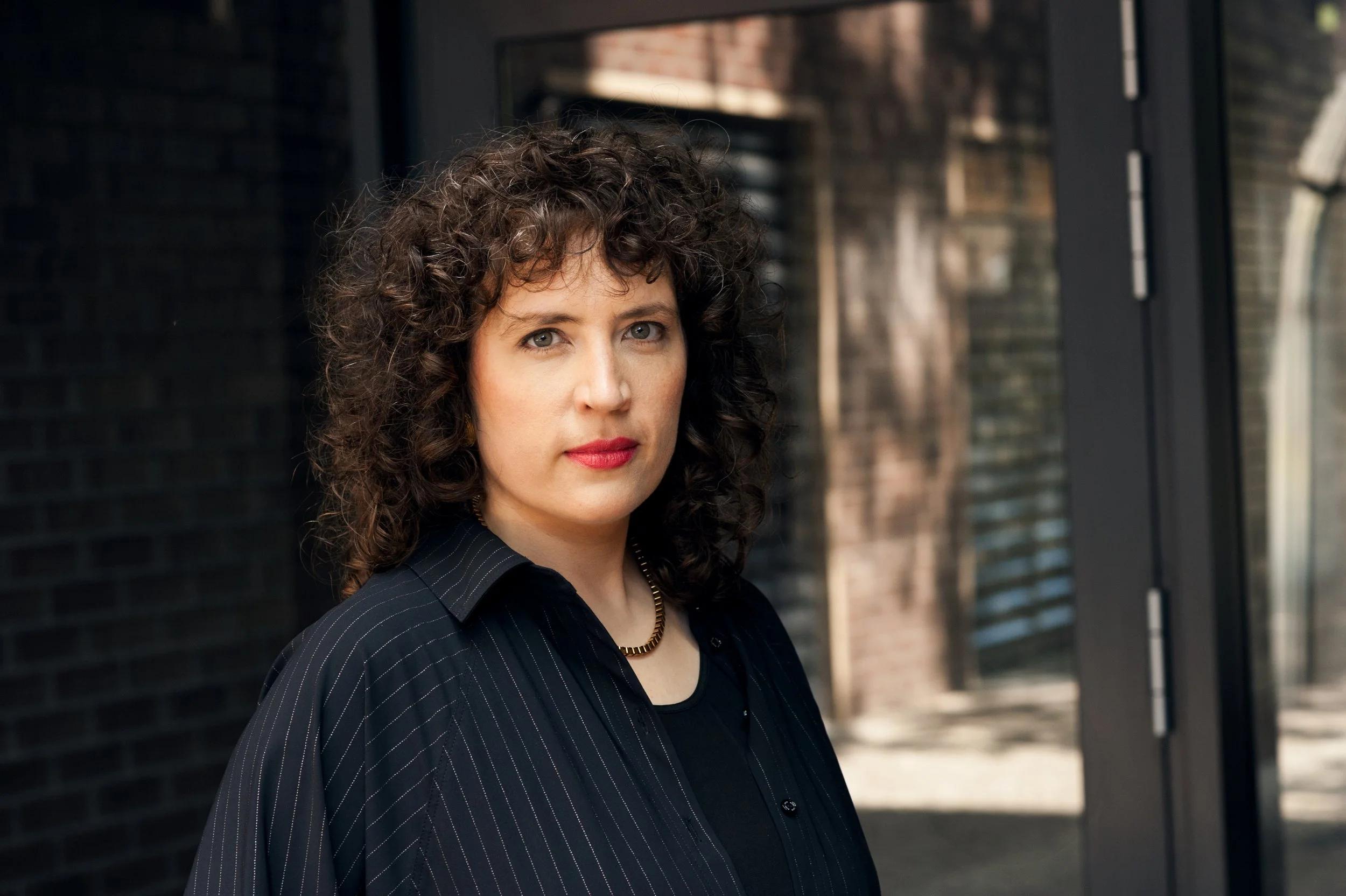 A woman with curly brown hair and red lipstick standing outdoors near a brick wall and black window frame.