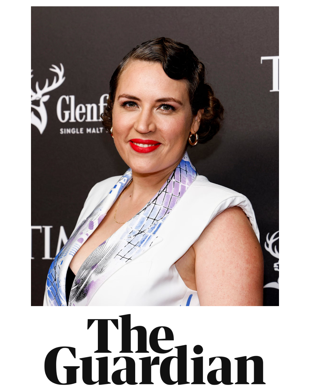 A woman with dark wavy hair styled in vintage waves, wearing a white sleeveless dress with a colorful patterned scarf, red lipstick, and gold hoop earrings, standing in front of a black backdrop with a logo for Glenfiddich.