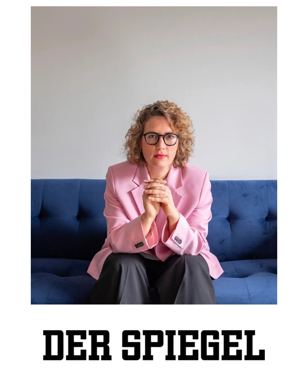 A woman with curly blonde hair and glasses wearing a pink blazer sitting on a blue sofa with her hands clasped together, looking directly at the camera against a plain white background.