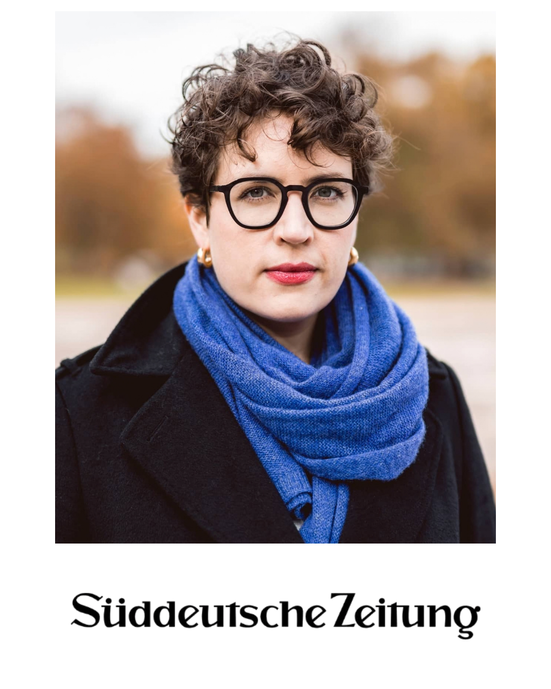 Portrait of a woman with short curly hair and glasses, wearing a black coat and a blue scarf, outdoors with blurred trees in the background.
