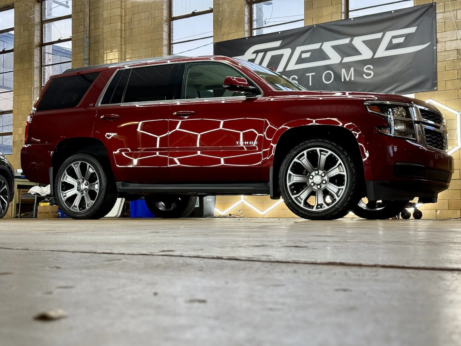 A red Chevrolet Tahoe SUV is parked indoors with a black sign behind it reading "GOSSE AUTOS". The SUV has chrome wheels and a shiny exterior reflecting the room's lights.