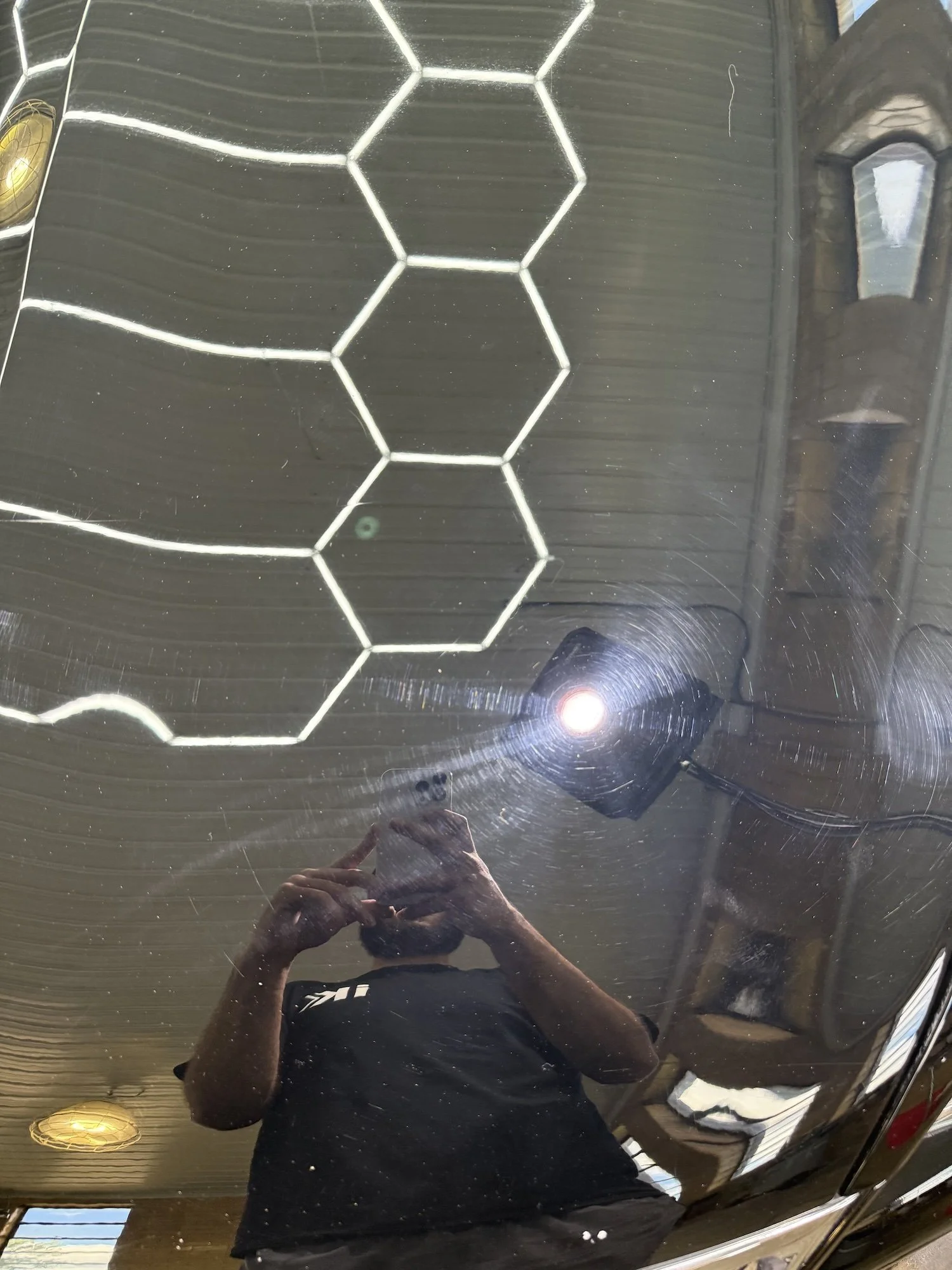 Close-up of the hood of a black sports car with visible scratches and reflections of overhead lights in a garage or workshop.