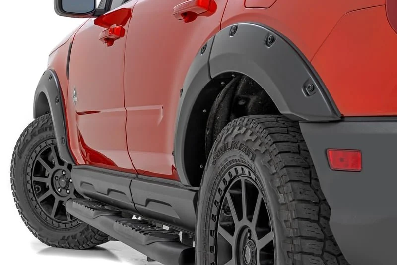 Close-up of a red off-road vehicle, showing wheel arches with black fender flares, large rugged tires, and a side step. The vehicle has a sporty, rugged design.