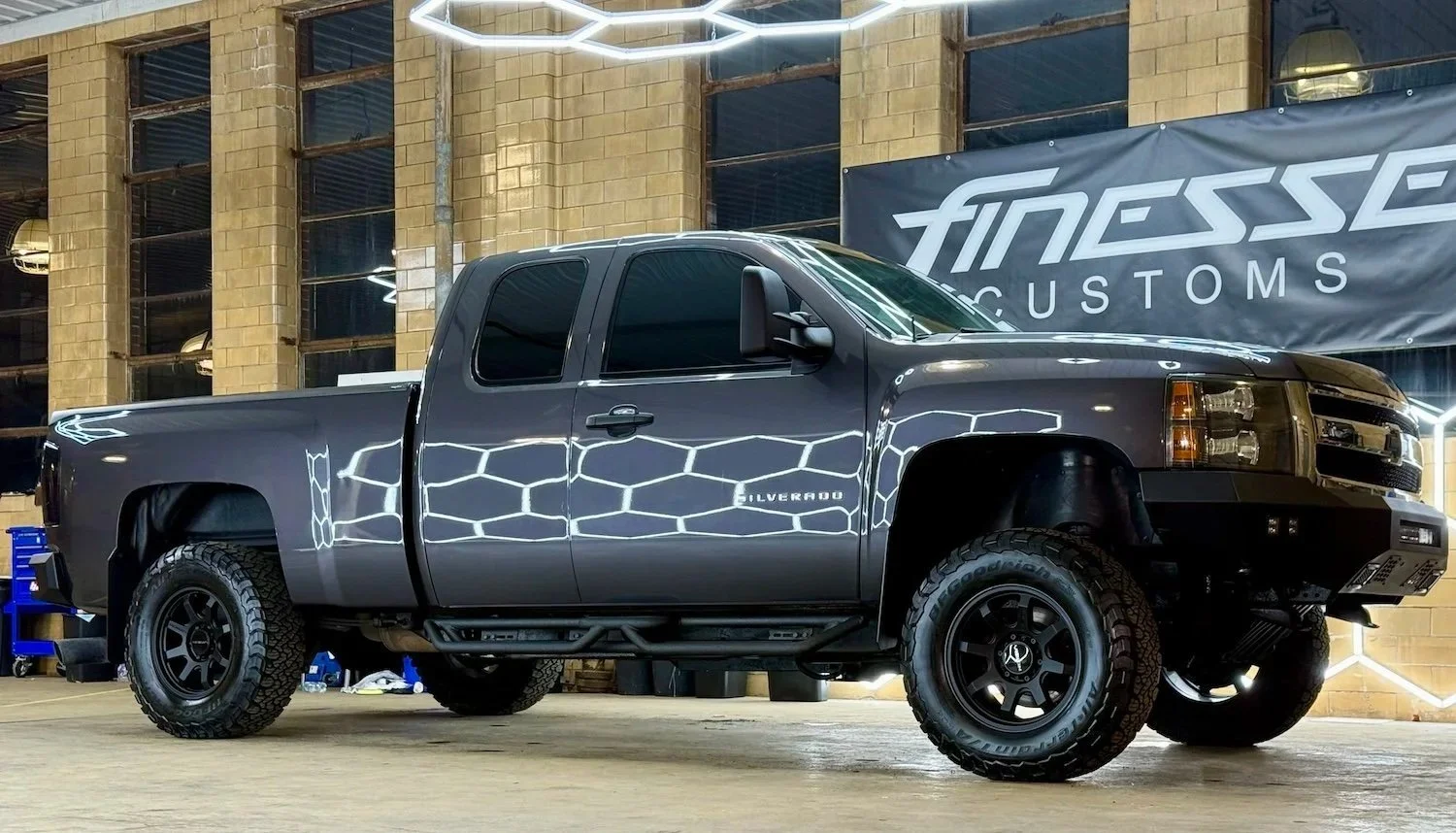 Black Chevrolet Silverado pickup truck with matte black wheels and off-road tires parked indoors with a backdrop of brick walls and a large banner that reads "Finess CUSTOMS".