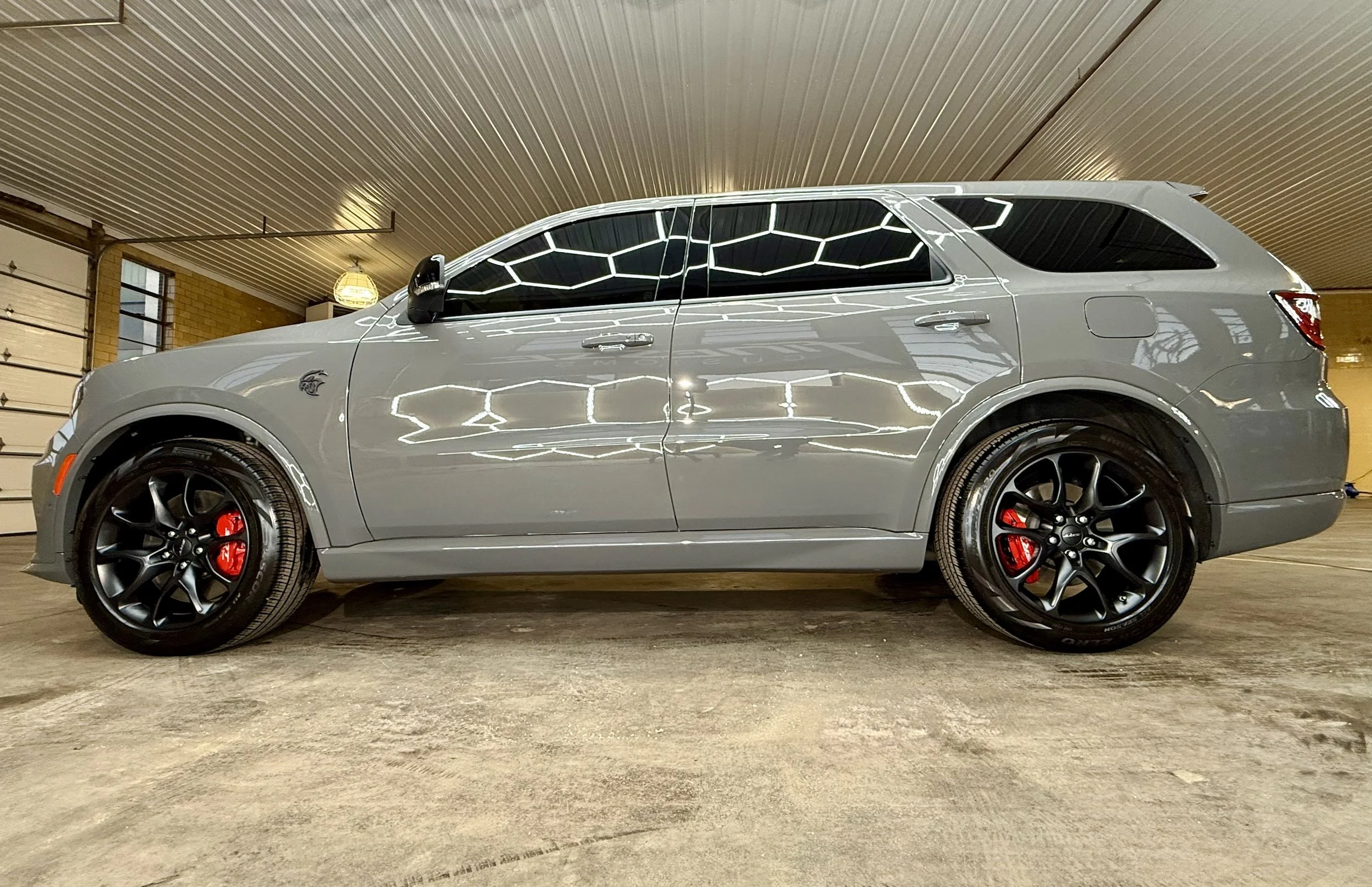A gray SUV with black wheels and red brake calipers parked inside a garage with a brick wall and a corrugated metal ceiling.