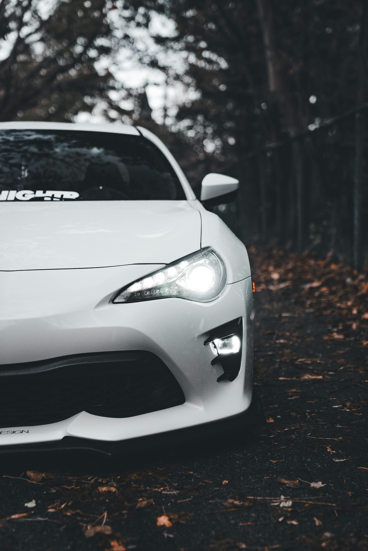 Front of a white modern sports car with headlights on, parked on a leaf-covered ground in a wooded area during dusk or dawn.
