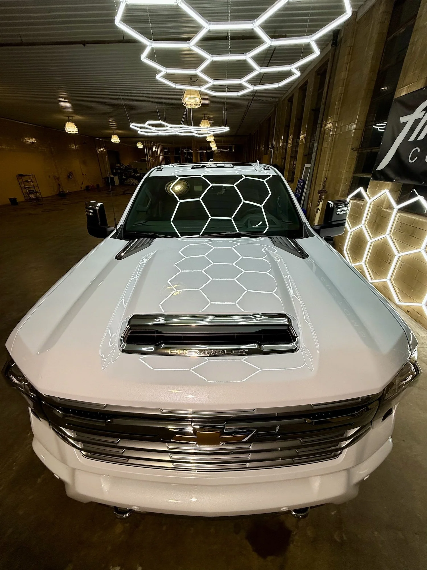 White Chevrolet pickup truck parked indoors under hexagon-shaped LED light fixtures, with honeycomb pattern reflections on the windshield and hood.