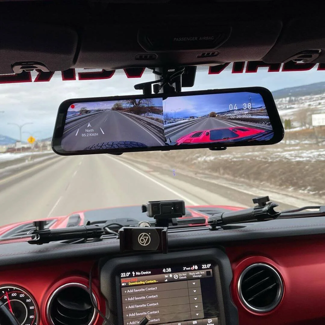 View from inside a vehicle showing a digital rearview mirror displaying a split screen with a front camera on left and rear camera on right, dashboard with a phone and other devices, and a road with snow-covered landscape ahead.