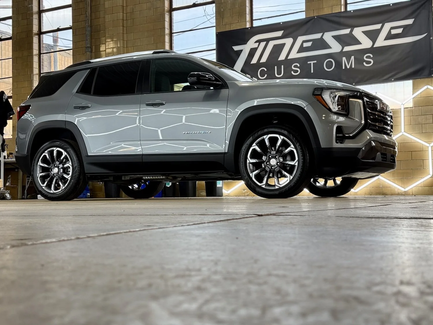 A silver GMC Terrain SUV parked indoors at a custom car shop with a "Finesse Customs" banner in the background.