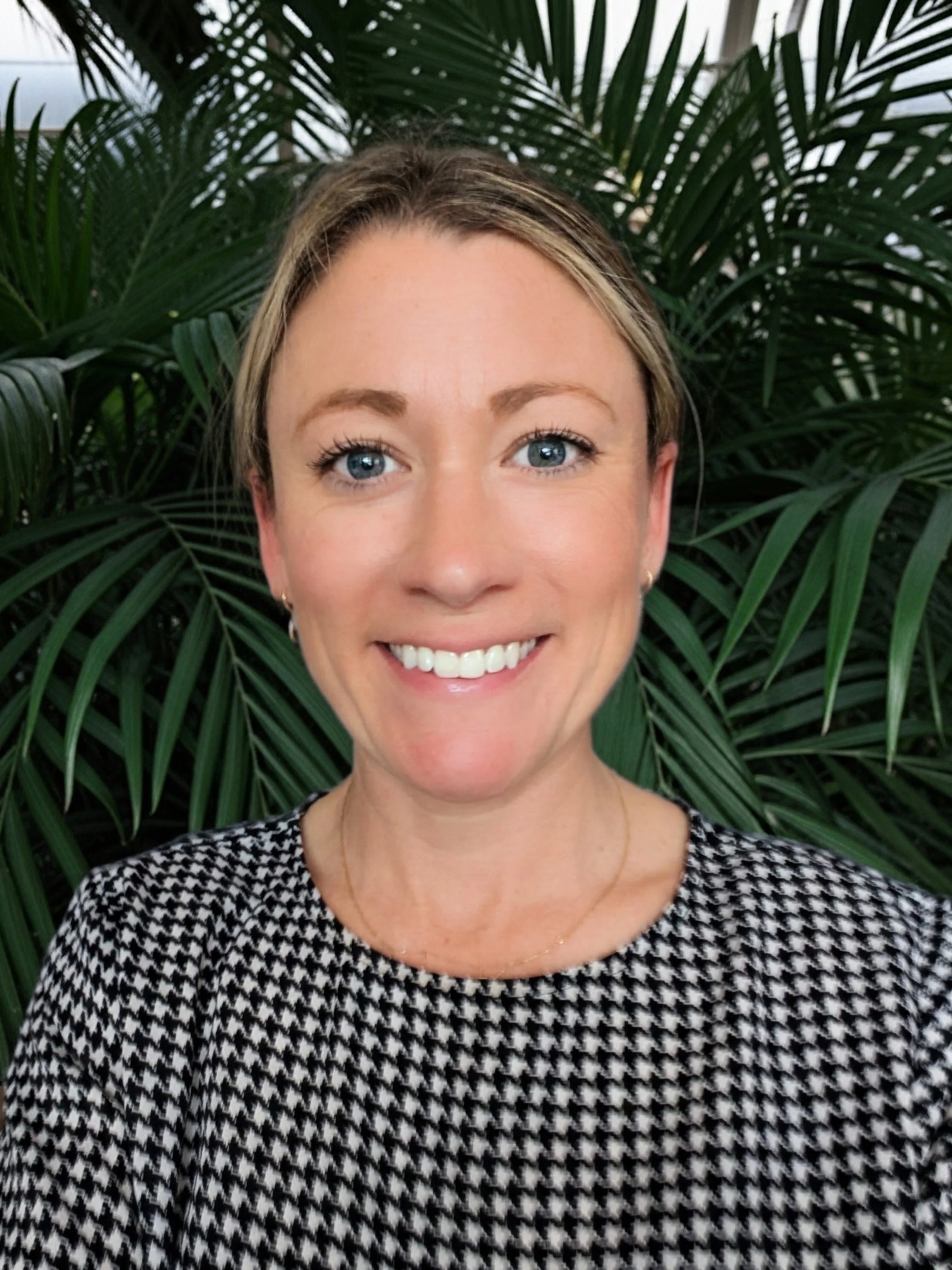 Close-up of a woman with blonde hair and blue eyes smiling, wearing a black and white houndstooth patterned shirt, with green palm leaves in the background.