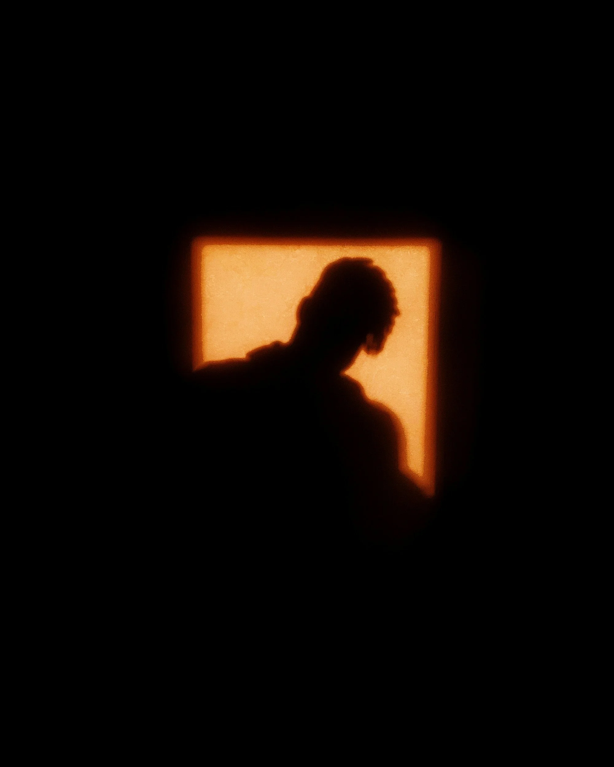 Silhouette of a person with dreadlocks, standing in front of a glowing orange window with a black background.