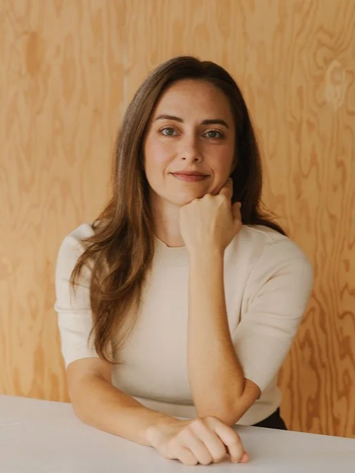 Portrait of Sarah with long brown hair sitting at a table, resting her chin on her right hand, smiling slightly, in front of a wooden wall.