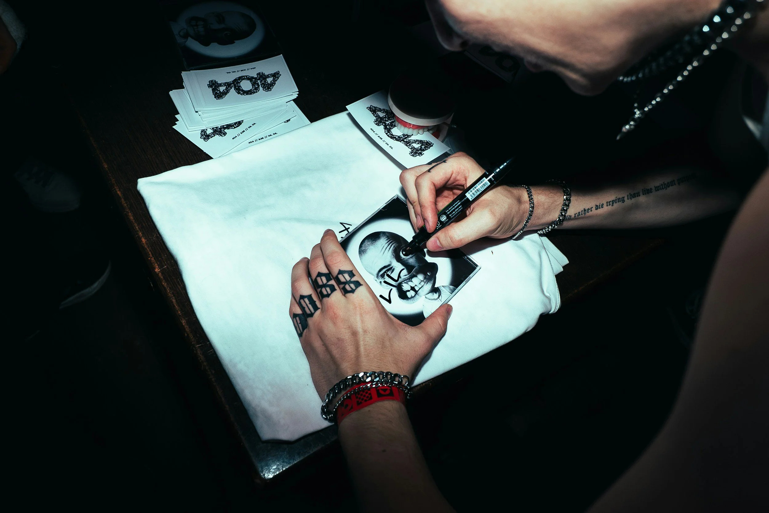 Person signing a photograph of a man with a shaved head and a big smile, at a table with stacks of stickers or photographs featuring the same man and the word 'AOA', and a white cloth.