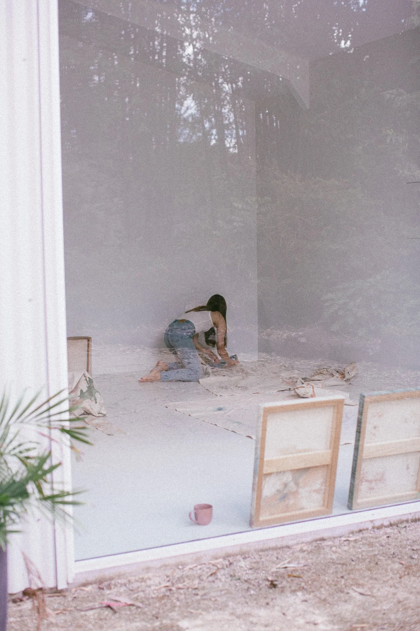 A woman in denim jeans and a white top is kneeling on the floor, painting or decorating an art piece in a bright, airy room with large windows showing trees outside. Two wooden chairs and a pink mug are visible in the foreground.