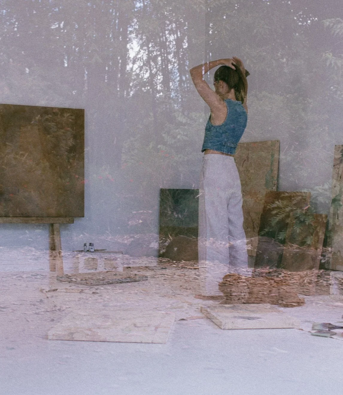 Reflection of a woman in an art studio, standing with arms raised, surrounded by paintings, art supplies, and a window with trees outside.