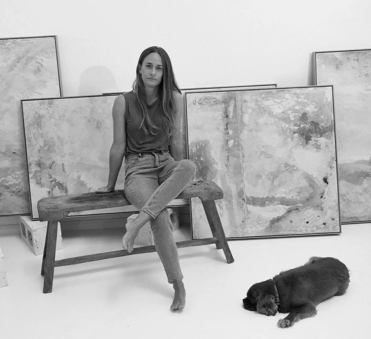 A woman sitting on a wooden bench in front of abstract paintings, with a puppy lying on the floor beside her, all in a minimalist art studio setting.