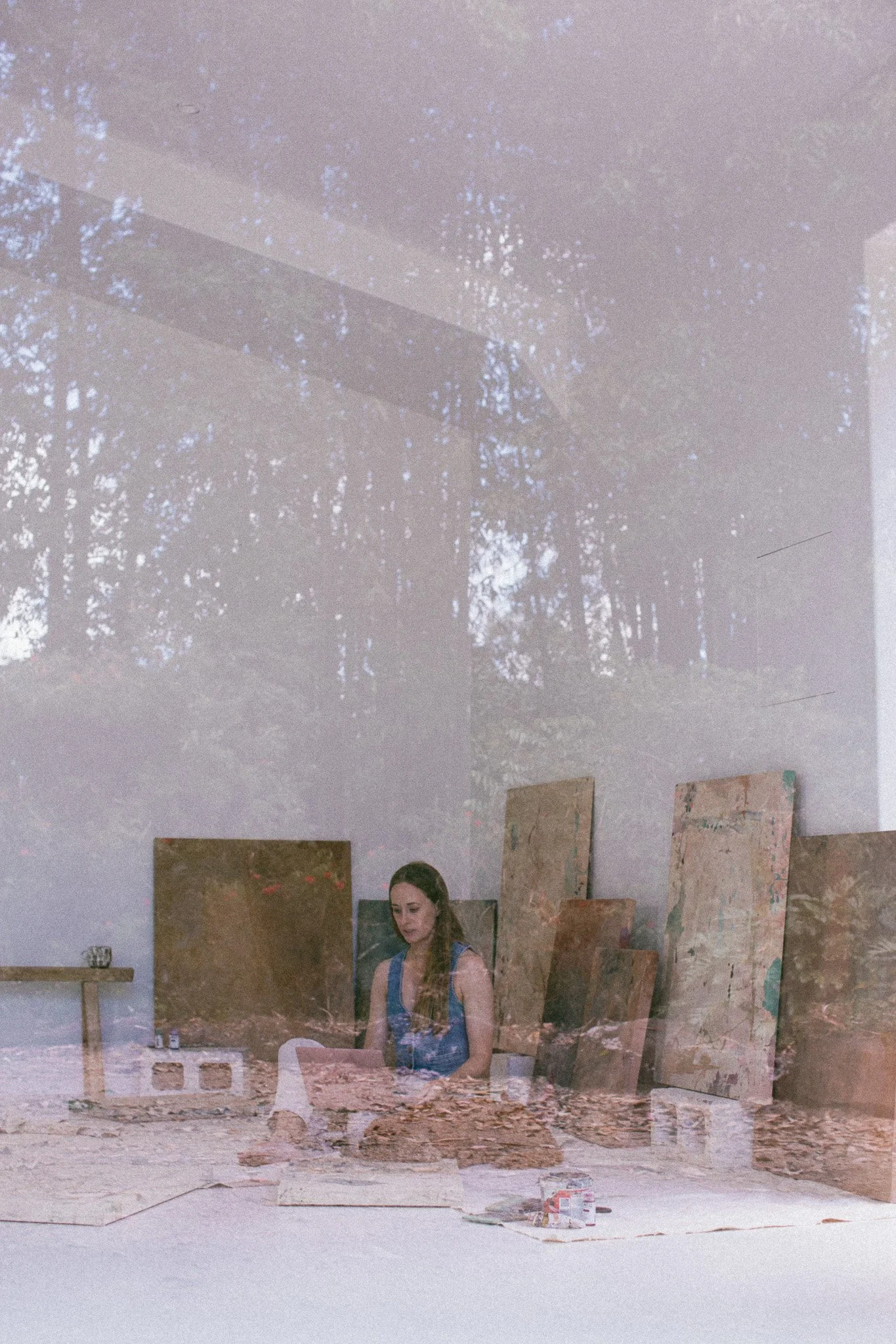 A woman sits on the floor of an art studio surrounded by completed paintings and art supplies. He is focusing on her artwork, with a shadowy tree reflected on the window.