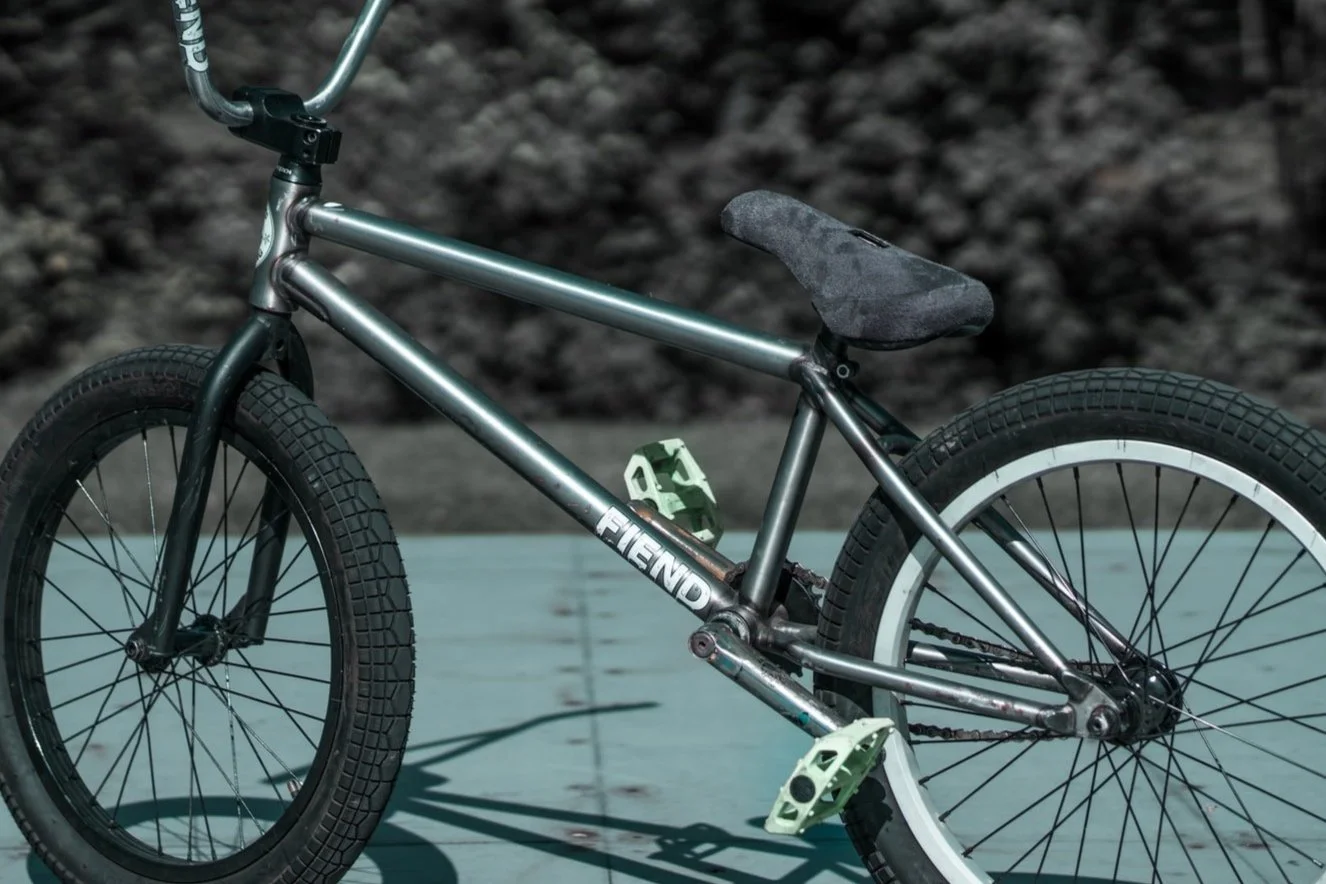 A diamond frame BMX bike with a black and silver frame, black tires, a black seat, green pedals, and a green water bottle holder, on a wooden surface outdoors.