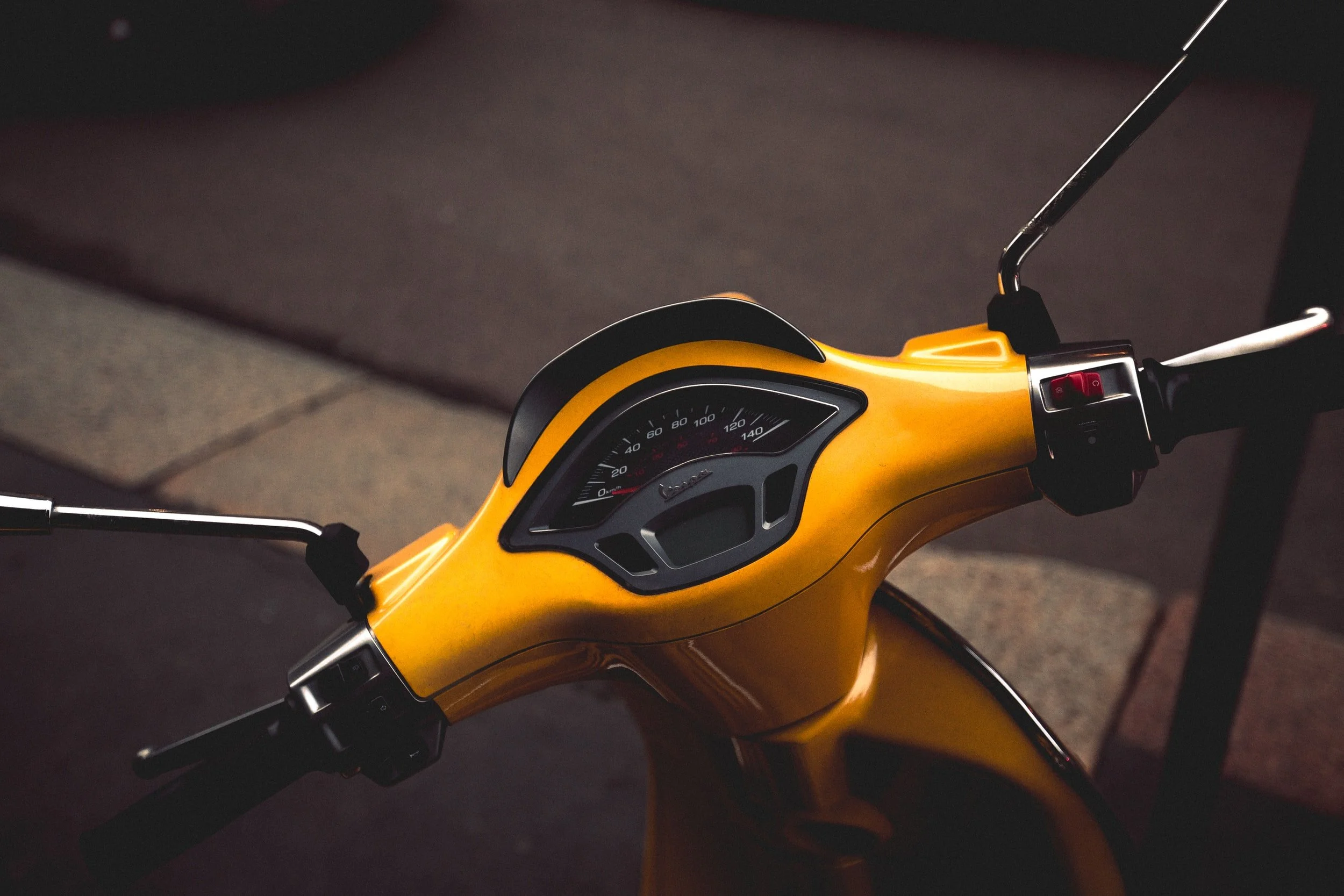 Close-up of a yellow Vespa scooter's handlebar and dashboard, with a speedometer gauge, set against a dark background.