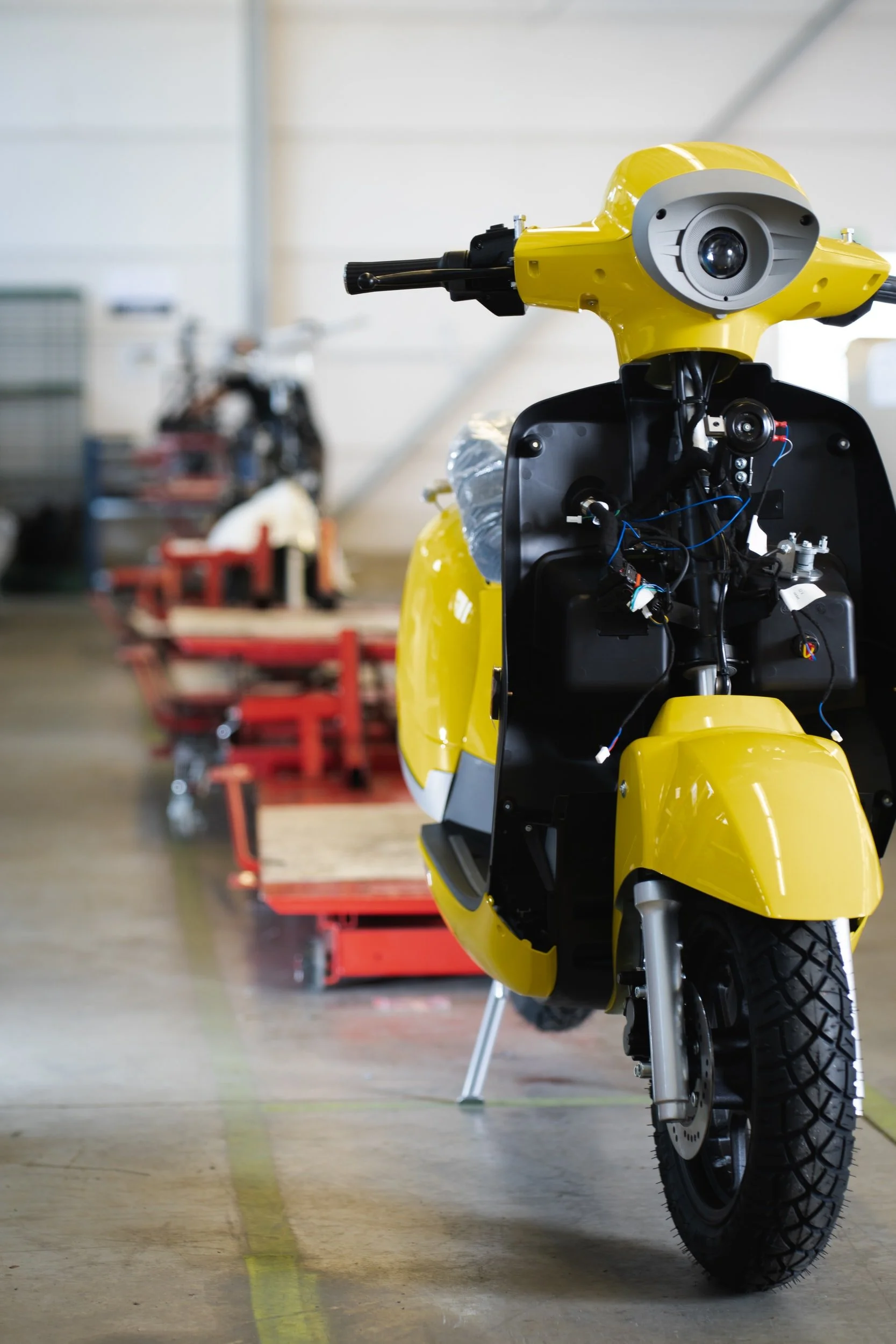 A yellow motor scooter, partially disassembled, inside a workshop with other similar vehicles in the background.