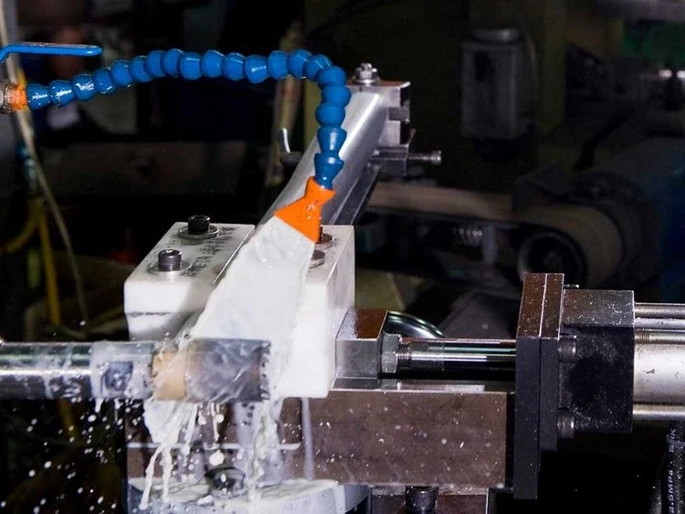 Close-up of a manufacturing machine with fluid being poured or sprayed, with blue flexible tubing connected to the machine in an industrial setting.