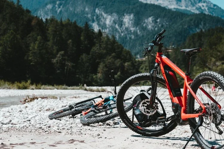 Red mountain bike with black front suspension fork lying on rocky riverbank, with two other bikes in the sand nearby, against a backdrop of trees and mountain ranges.
