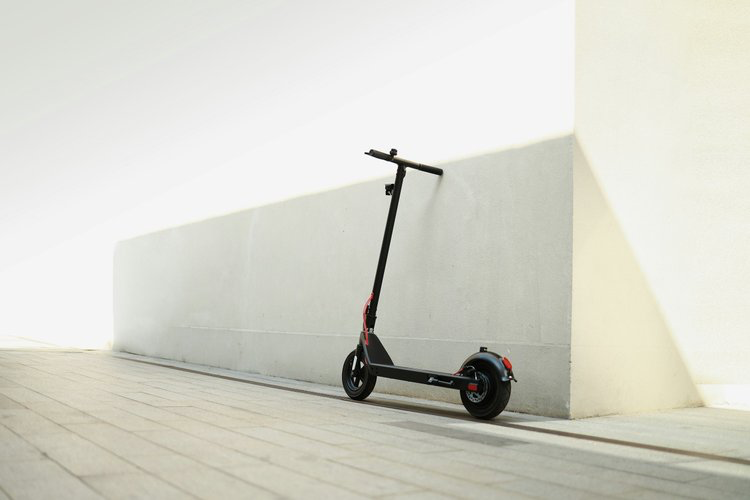 Black electric scooter leaning against a white wall outdoors on a paved surface.